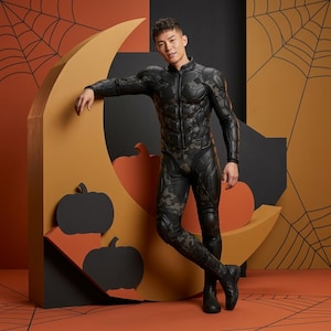 Könnte beinhalten: Ein schwarzer und camouflagefarbener Overall mit hohem Kragen und Reißverschluss. Das Kostüm hat gepolsterte Details und wird mit schwarzen Stiefeln getragen. Der Hintergrund zeigt Halloween-Dekorationen wie Kürbisse und Spinnweben.
