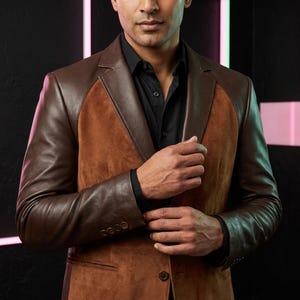 May include: A brown blazer featuring a combination of smooth leather sleeves and a suede front panel. The garment includes a notched lapel, a single button closure, and four small buttons on each cuff, worn over a solid black button-down shirt.