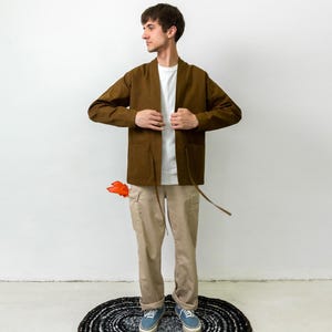 Puede incluir: Una chaqueta marr&oacute;n abierta por delante con mangas largas y dos bolsillos, usada sobre una camiseta blanca lisa. La chaqueta cuenta con finas cintas marrones en la cintura. El conjunto se completa con pantalones cargo beige y zapatillas azules.