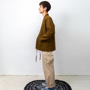 Puede incluir: Una chaqueta de manga larga color verde oliva con cuello en V y cierre de lazo lateral, combinada con pantalones cargo beige con bolsillos laterales y zapatillas de lona azules. El conjunto lo lleva una persona de perfil ante una pared.