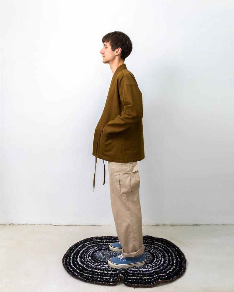 Puede incluir: Una chaqueta de manga larga color verde oliva con cuello en V y cierre de lazo lateral, combinada con pantalones cargo beige con bolsillos laterales y zapatillas de lona azules. El conjunto lo lleva una persona de perfil ante una pared.