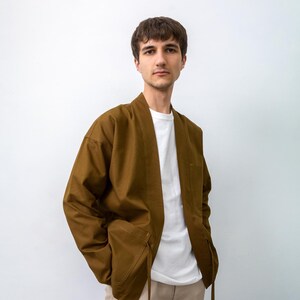 Puede incluir: Una chaqueta marr&oacute;n abierta por delante con mangas largas y cuello en V, que presenta dos bolsillos delanteros con tiras de tela colgantes. La chaqueta se lleva sobre una camiseta blanca lisa de cuello redondo y pantalones cargo beige claro.
