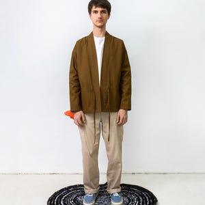 Puede incluir: Una chaqueta marr&oacute;n abierta por delante con mangas largas y finas cintas en la cintura, sobre una camiseta blanca lisa. El conjunto incluye pantalones de color beige claro y zapatillas de lona azules, con un objeto naranja parcialmente visible.
