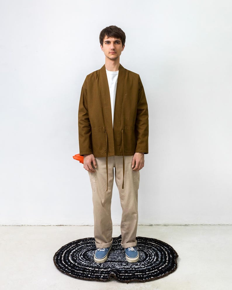 Puede incluir: Una chaqueta marr&oacute;n abierta por delante con mangas largas y finas cintas en la cintura, sobre una camiseta blanca lisa. El conjunto incluye pantalones de color beige claro y zapatillas de lona azules, con un objeto naranja parcialmente visible.