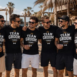 May include: Six black t-shirts featuring white text that reads TEAM GROOM, the name THOMAS, and the date EVG 2025. Each shirt includes a gold crown graphic above the text and a small white bow tie graphic below the name, worn by six individuals.
