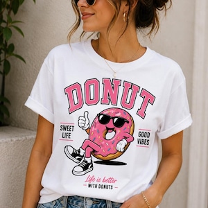 May include: A white short-sleeve t-shirt featuring a graphic of a pink frosted donut wearing black sunglasses and sneakers. The text reads DONUT, SWEET LIFE, GOOD VIBES, and Life is better WITH DONUTS in pink and black lettering on the front.