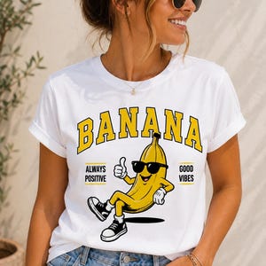 May include: A white short-sleeve t-shirt featuring a graphic of a yellow banana wearing black sunglasses and black sneakers. The banana is giving a thumbs up. The text BANANA is printed in yellow above, with ALWAYS POSITIVE GOOD VIBES on sides.