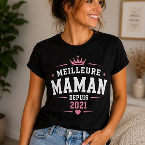 Puede incluir: Una camiseta negra de manga corta con un estampado gr&aacute;fico en blanco y rosa. El dise&ntilde;o incluye una corona rosa, el texto "MEILLEURE MAMAN DEPUIS 2021" en letras blancas y rosas, y un peque&ntilde;o coraz&oacute;n rosa en la parte inferior del gr&aacute;fico.
