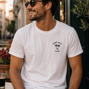 May include: A white short-sleeved t-shirt featuring a small black graphic on the left chest. The graphic consists of two palm trees with the text "GOOD DAYS" arched above and the word "CLUB" positioned directly below the palm trees.