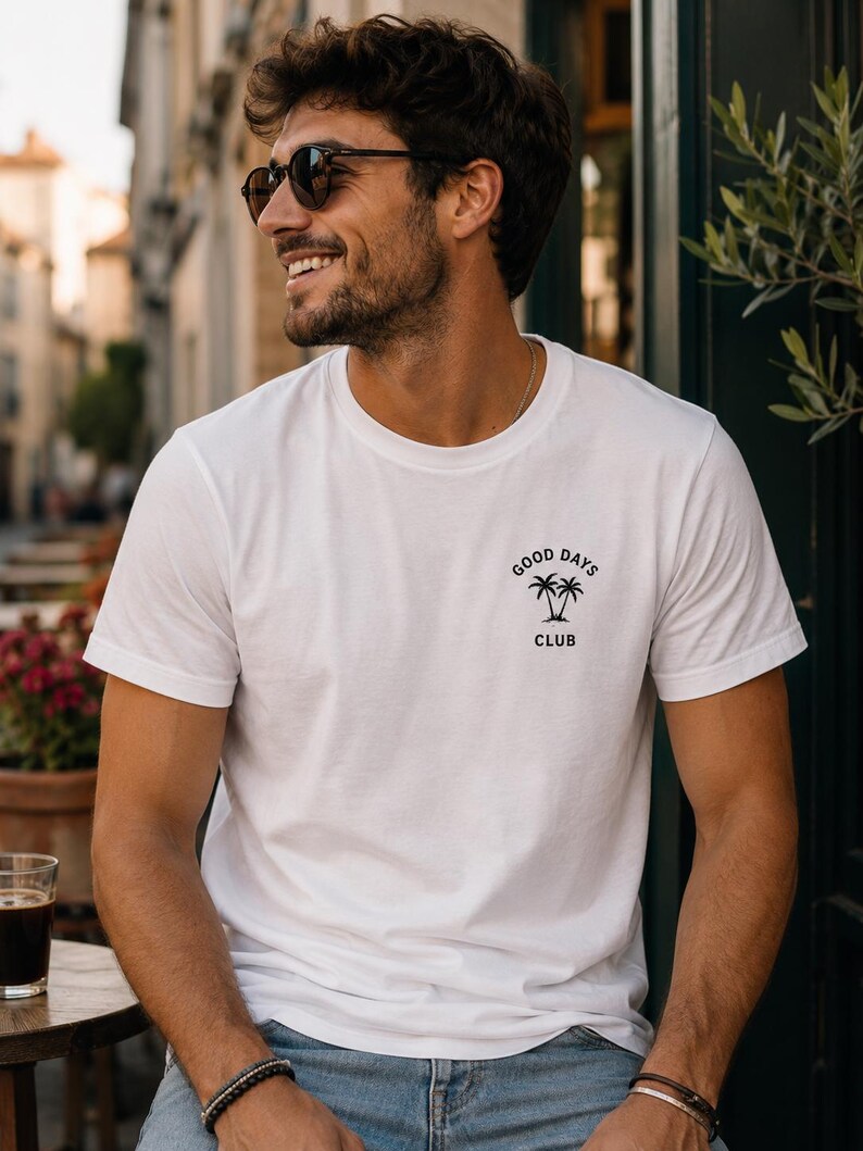 May include: A white short-sleeved t-shirt featuring a small black graphic on the left chest. The graphic consists of two palm trees with the text "GOOD DAYS" arched above and the word "CLUB" positioned directly below the palm trees.