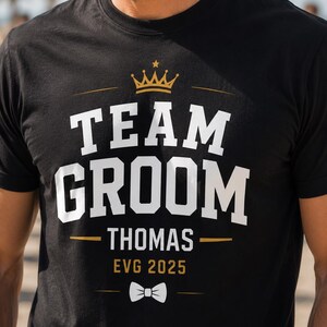 May include: A black short-sleeve t-shirt featuring a gold crown graphic above the white text "TEAM GROOM" and "THOMAS" in a bold font. Below the name, the text "EVG 2025" appears in gold, followed by a small white bow tie graphic.
