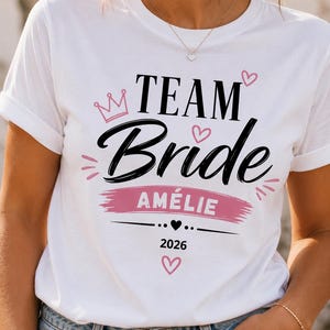 May include: A white t-shirt featuring the black text "TEAM Bride" with a pink crown and heart accents. Below, a pink brushstroke background highlights the name "AM&Eacute;LIE" in white, followed by a small black heart and the year "2026" in black text.