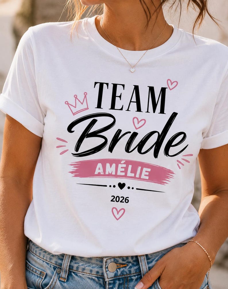 May include: A white t-shirt featuring the black text "TEAM Bride" with a pink crown and heart accents. Below, a pink brushstroke background highlights the name "AM&Eacute;LIE" in white, followed by a small black heart and the year "2026" in black text.
