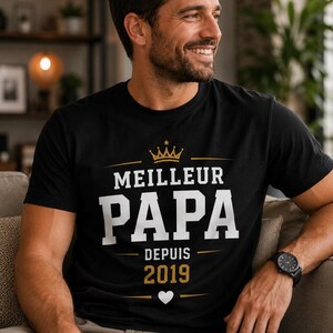 May include: A black short-sleeved t-shirt featuring a gold crown graphic above the white text "MEILLEUR PAPA" and the gold text "DEPUIS 2019" with a small white heart icon centered below the year, all printed on the front of the garment.