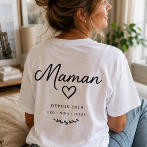 May include: A white short-sleeved t-shirt featuring black text on the back that reads Maman, a heart icon, the text DEPUIS 2019, the names L&Eacute;O, NINA, and JULES, and a small decorative branch graphic at the bottom of the printed design.