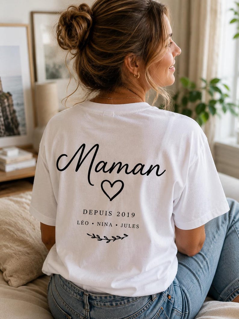 May include: A white short-sleeved t-shirt featuring black text on the back that reads Maman, a heart icon, the text DEPUIS 2019, the names L&Eacute;O, NINA, and JULES, and a small decorative branch graphic at the bottom of the printed design.