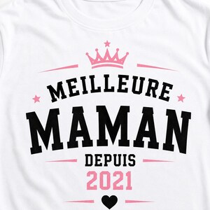 Puede incluir: Una camiseta blanca con un dise&ntilde;o gr&aacute;fico que incluye una corona rosa, estrellas rosas y un coraz&oacute;n negro. El texto MEILLEURE MAMAN DEPUIS 2021 aparece en letras negras y rosas llamativas, dispuestas en el centro de la prenda.