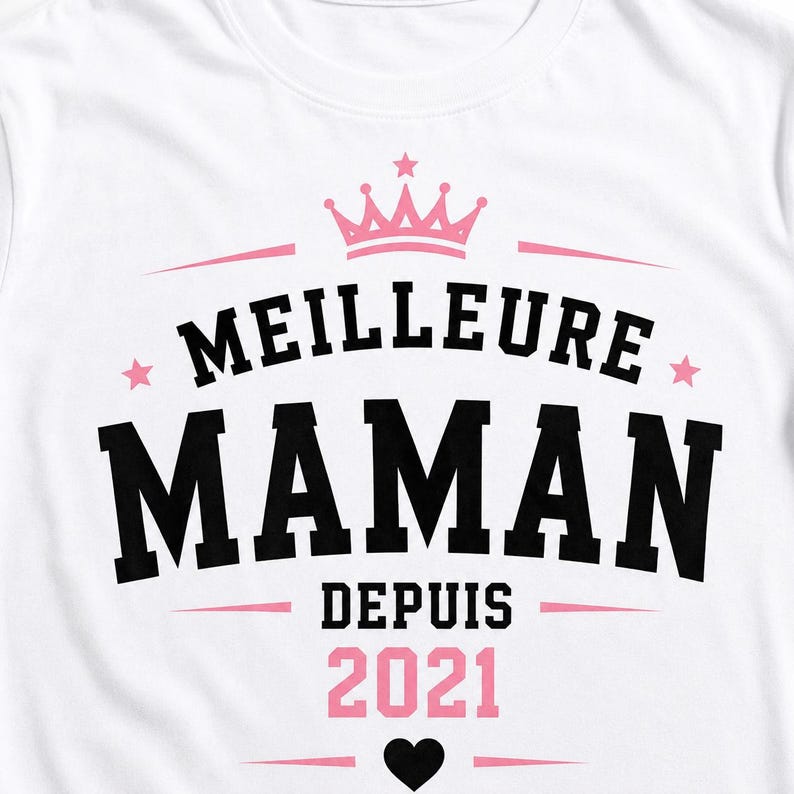 Puede incluir: Una camiseta blanca con un dise&ntilde;o gr&aacute;fico que incluye una corona rosa, estrellas rosas y un coraz&oacute;n negro. El texto MEILLEURE MAMAN DEPUIS 2021 aparece en letras negras y rosas llamativas, dispuestas en el centro de la prenda.