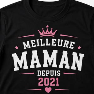 Puede incluir: Una camiseta negra con un dise&ntilde;o gr&aacute;fico que presenta una corona rosa en la parte superior, el texto blanco MEILLEURE MAMAN y el texto rosa DEPUIS 2021 debajo. El dise&ntilde;o incluye estrellas rosas a los lados y un peque&ntilde;o coraz&oacute;n rosa en el centro.