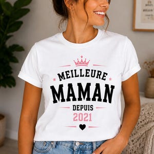 Puede incluir: Una camiseta blanca de manga corta con el texto MEILLEURE MAMAN DEPUIS 2021 en letras negras y rosas. El dise&ntilde;o incluye un icono de corona rosa sobre el texto, peque&ntilde;as estrellas rosas a los lados y un icono de coraz&oacute;n negro s&oacute;lido abajo.