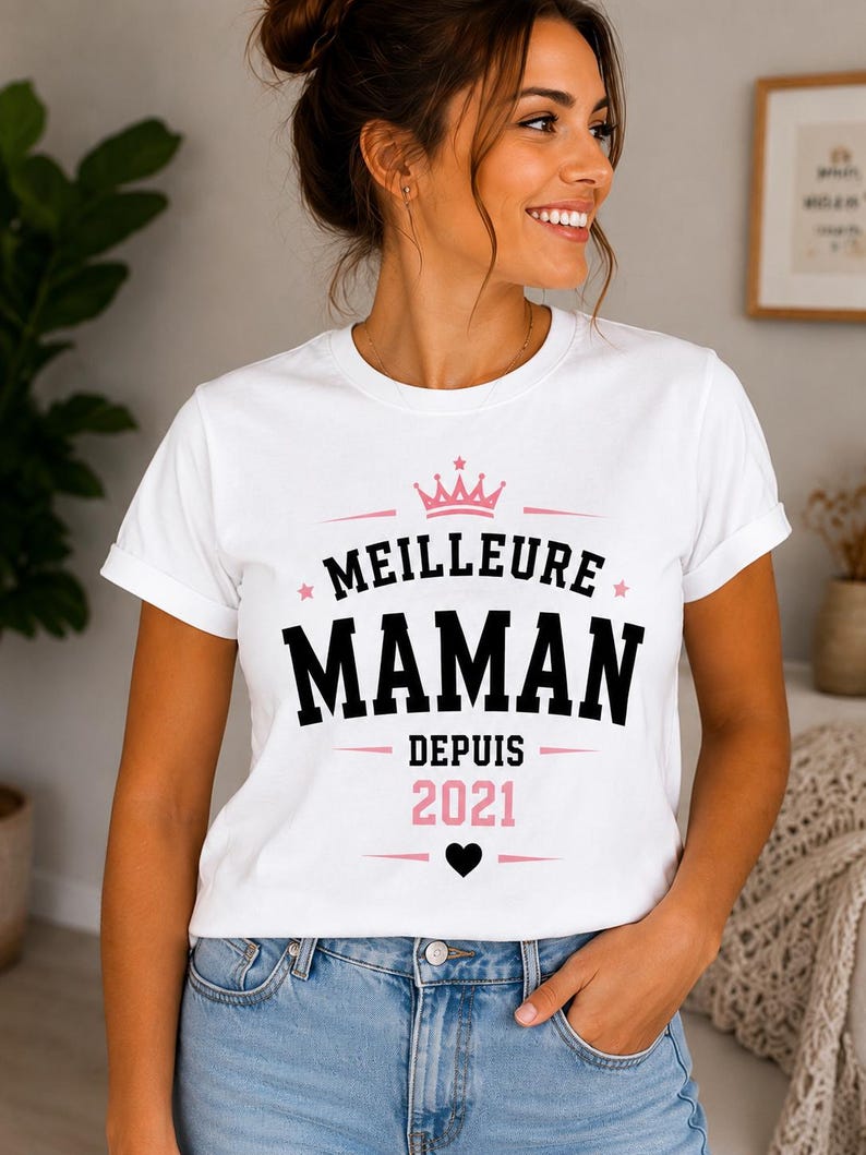 Puede incluir: Una camiseta blanca de manga corta con el texto MEILLEURE MAMAN DEPUIS 2021 en letras negras y rosas. El dise&ntilde;o incluye un icono de corona rosa sobre el texto, peque&ntilde;as estrellas rosas a los lados y un icono de coraz&oacute;n negro s&oacute;lido abajo.