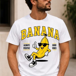 May include: A white t-shirt featuring a graphic of a yellow banana character wearing black sunglasses and sneakers. The banana is giving a thumbs up. The text BANANA is printed in yellow with a black outline above, and ALWAYS POSITIVE GOOD VIBES is printed in black text on either side.
