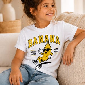 K&ouml;nnte beinhalten: Ein wei&szlig;es Kurzarm-T-Shirt mit einer Grafik einer gelben Banane, die eine schwarze Sonnenbrille und schwarze Turnschuhe tr&auml;gt. Die Banane zeigt einen Daumen hoch. Der Text BANANA steht in Gelb dar&uuml;ber, mit ALWAYS POSITIVE und GOOD VIBES an den Seiten.