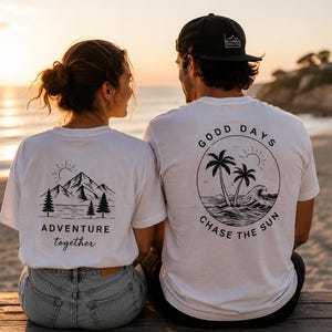 May include: Two white t-shirts featuring black graphic prints on the back. One shirt displays a mountain, trees, and the text "ADVENTURE together." The other shirt shows palm trees, a wave, and the text "GOOD DAYS CHASE THE SUN."