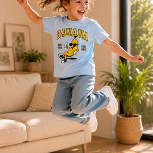 K&ouml;nnte beinhalten: Ein hellblaues kurz&auml;rmeliges T-Shirt mit einer Grafik einer Cartoon-Banane, die eine Sonnenbrille und Turnschuhe tr&auml;gt. Der Text BANANA ist in Gelb &uuml;ber der Figur gedruckt, mit den Worten ALWAYS POSITIVE und GOOD VIBES an den Seiten.
