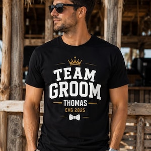 May include: A black short-sleeved t-shirt featuring a gold crown graphic above the white text "TEAM GROOM" and "THOMAS". Below the name, the text "EVG 2025" is printed in gold, followed by a small white bow tie graphic centered on the chest.