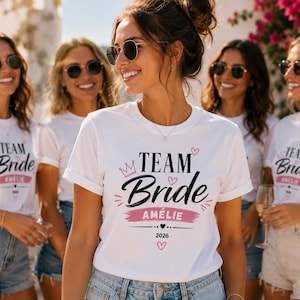 May include: A white crew neck t-shirt featuring the black text "TEAM Bride" with a pink crown icon, pink hearts, and a pink brush stroke graphic containing the name "AM&Eacute;LIE" in white letters, followed by the year "2026" and a small black heart.