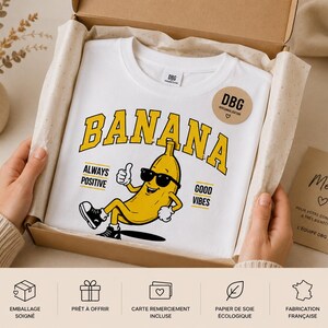 K&ouml;nnte beinhalten: Ein wei&szlig;es T-Shirt mit einer gelben Cartoon-Banane, die eine schwarze Sonnenbrille tr&auml;gt, einen Daumen hoch zeigt und sitzt. Der Text BANANA steht in gro&szlig;en gelben Buchstaben dar&uuml;ber, mit ALWAYS POSITIVE und GOOD VIBES an den Seiten.