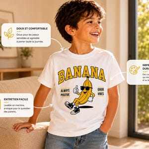 K&ouml;nnte beinhalten: Ein wei&szlig;es Kurzarm-T-Shirt mit einer Grafik einer gelben Bananenfigur, die eine Sonnenbrille und Turnschuhe tr&auml;gt und den Daumen hochh&auml;lt. Der Text BANANA steht in Gelb dar&uuml;ber, mit ALWAYS POSITIVE und GOOD VIBES an den Seiten.