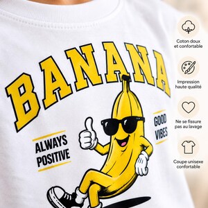 K&ouml;nnte beinhalten: Ein wei&szlig;es Sweatshirt mit Rundhalsausschnitt und einer gro&szlig;en gelb-schwarzen Grafik einer Cartoon-Banane mit Sonnenbrille und schwarzen Turnschuhen, die den Daumen hochh&auml;lt. Der Text lautet BANANA, ALWAYS POSITIVE und GOOD VIBES.