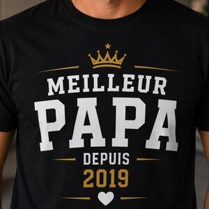 May include: A black short-sleeved t-shirt featuring a graphic print with a gold crown above the white text MEILLEUR PAPA. Below this, the text DEPUIS 2019 appears in gold, followed by a small white heart centered between two horizontal gold lines.