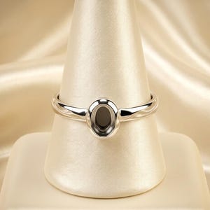 以下が含まれることがあります： クリーム色の背景に、白いスタンドに展示された、楕円形のセッティングが施された磨かれたシルバーリング。 リングのバンドは滑らかで丸みを帯びており、セッティングは宝石を保持するように設計されています。 リングはジュエリーです。