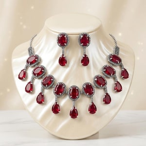 Può includere: Un set di gioielli abbagliante con collana e orecchini abbinati. Il set presenta grandi pietre preziose rosse scure a taglio ovale, accentate da pietre più piccole e trasparenti. La collana e gli orecchini sono esposti su un busto bianco, su uno sfondo morbido e sfocato.