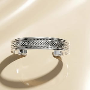 Op de afbeelding: Een zilveren manchetarmband met een geweven touwontwerp. De open manchet heeft een textuurpatroon rond de band. De armband is gemaakt van gepolijst metaal en wordt gepresenteerd op een lichtbeige oppervlak.