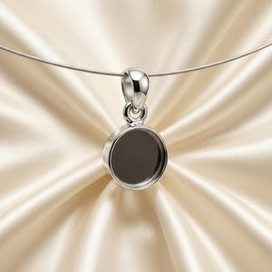 May include: A silver pendant with a circular setting and a loop for a chain. The pendant has a dark, recessed center and hangs from a thin, silver chain. The background is a soft, cream-colored fabric.