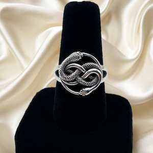 May include: A silver-toned ring featuring a circular design with a detailed snake coiled into an infinity shape. The snake has textured scales and is positioned within a smooth, polished circular frame that connects to a solid, plain band.