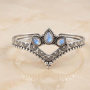 Può includere: Un bracciale argentato con un design a forma di corona. Il bracciale presenta tre pietre iridescenti a forma di goccia e dettagli intricati. La fascia è adornata da piccoli abbellimenti circolari e un motivo ripetuto.
