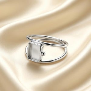 May include: A silver-toned ring featuring a double band design that connects to a central rectangular bezel setting. The bezel is empty and has a smooth, polished finish. A small, spherical metal accent is attached to the side of the rectangle.