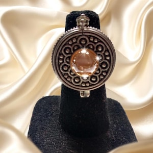 Könnte beinhalten: Ein kreisförmiger Ring in Silberoptik mit einem verzierten, geprägten Metallrand und kleinen perlenartigen Details, die einen zentralen, glatten, durchscheinenden bernsteinfarbenen Cabochon-Stein umschließen, der das Licht reflektiert.