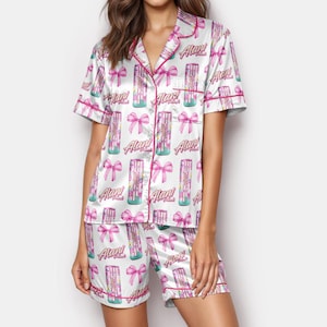 Puede incluir: Conjunto de pijama de satén blanco con un estampado repetido de lazos rosas y latas de bebidas con el texto "Alan!". El conjunto incluye una camisa de manga corta con botones y pantalones cortos a juego, ambos ribeteados con ribete rosa.