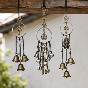 May include: Three hanging ornaments featuring silver-toned rings, black cords, and bronze-colored metal charms. The charms include pentagrams, triquetra knots, ornate keys, small bells, and a seated deity figure, all suspended from a wooden beam.
