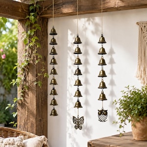 May include: Three vertical hanging wind chimes made of antique brass-colored metal bells. Each strand features a series of small, uniform bells suspended on a thin chain. The center strand ends with a butterfly charm, and the right strand ends with an owl charm.