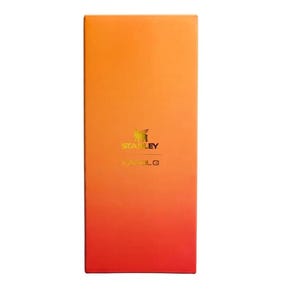 May include: A rectangular product box featuring a vertical gradient that transitions from orange at the top to red at the bottom. The box displays the gold Stanley logo and the text KAROL G printed in a gold font centered on the front surface.