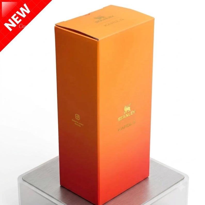 May include: A rectangular cardboard product box featuring a vertical gradient color scheme that transitions from orange at the top to red at the bottom. The box displays the gold Stanley logo and text on the front and top, with a small Instagram icon.