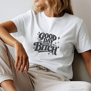 Könnte beinhalten: Ein weißes Kurzarm-T-Shirt mit dunkelgrauem Text auf der Brust, der GOOD DAY TO BE A BITCH lautet, begleitet von kleinen Sternsymbolen. Das T-Shirt hat einen Rundhalsausschnitt und wird mit einer hellen Jogginghose mit Kordelzug kombiniert.