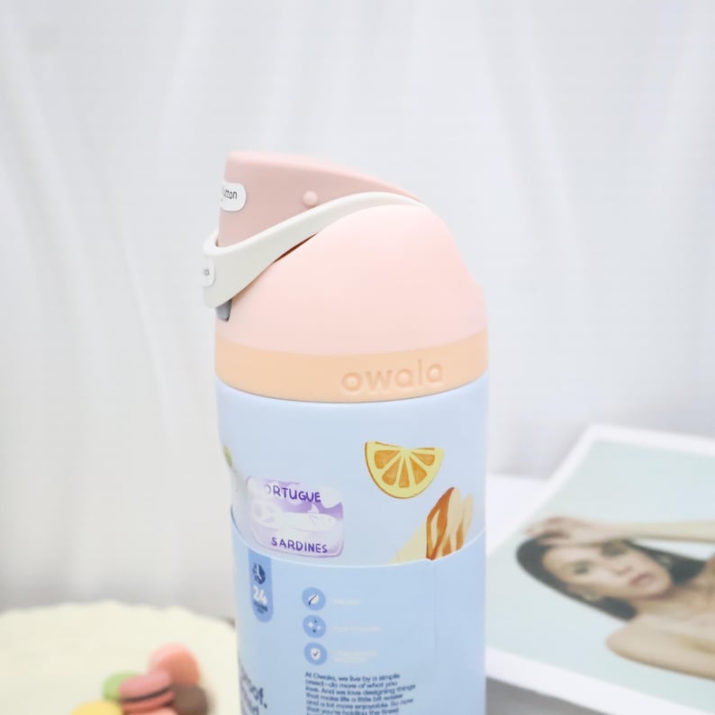 May include: A light blue Owala water bottle with a pink lid and a white handle. The bottle has a design with the words "PORTUGUE SARDINES" and a lemon slice illustration. The bottle is made of plastic.